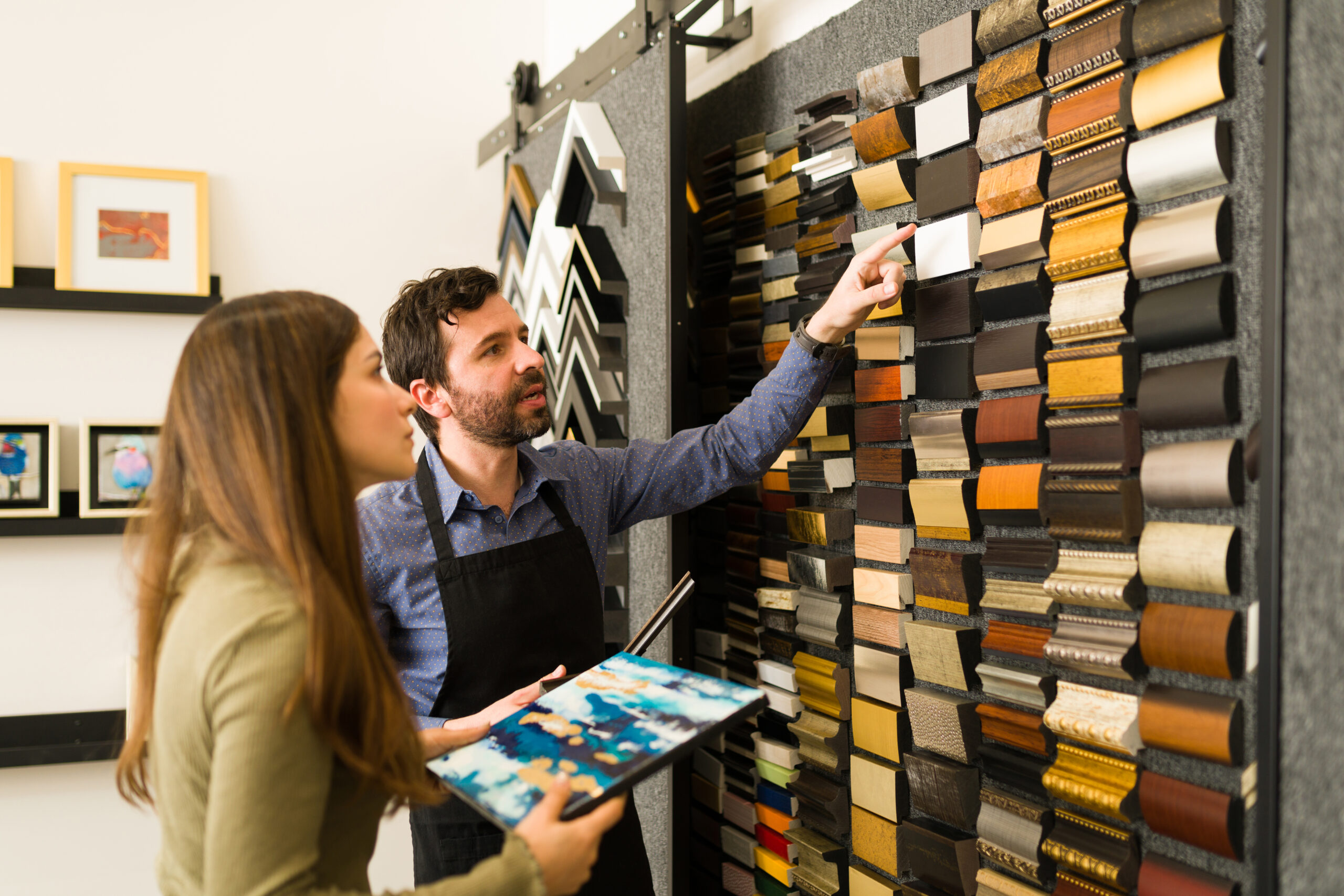Attractive salesman and pretty woman in her 20s are in a store and looking at a wall full of frame samples